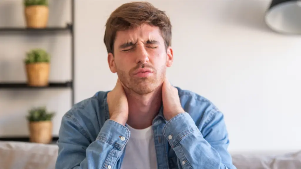 A imagem mostra um homem sentado em um sofá com os olhos fechados e as mãos sobre a nuca. Seu rosto está aflito, pois está sentindo dores na região do pescoço.