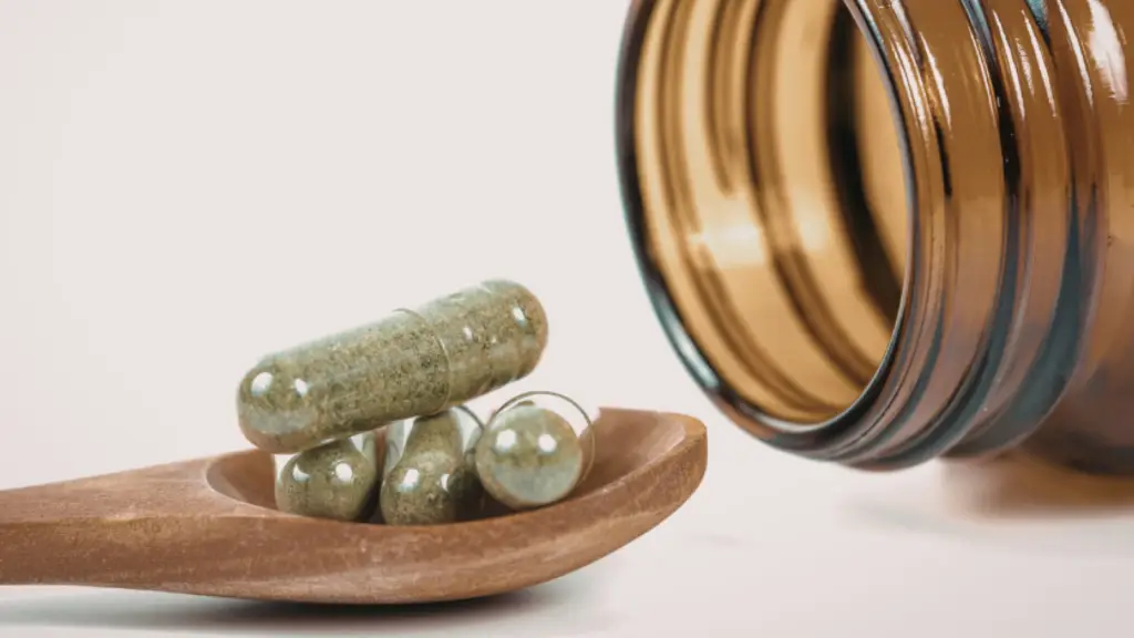 A imagem mostra, ao lado esquerdo, uma colher de madeira com quatro cápsulas de suplementos alimentares. Já ao lado direito, tem um pote de suplemento aberto e deitado, que é de onde saíram as cápsulas.