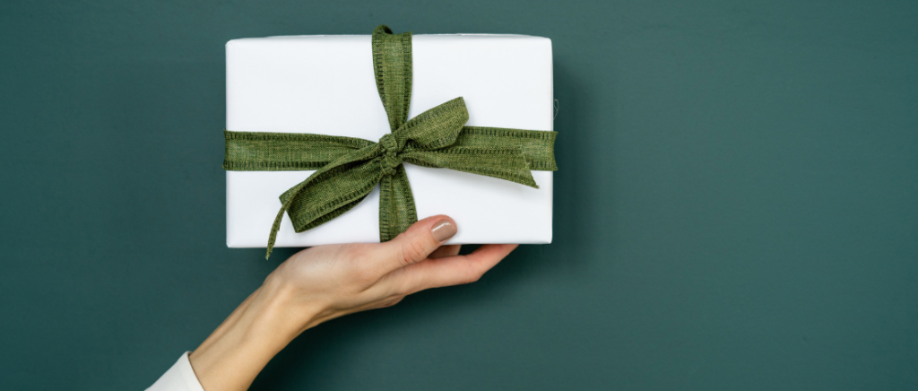A imagem mostra a mão de uma pessoa segurando uma caixa de presente na cor branca com um laço verde, representando uma pessoa que quer dar um presente para quem gosta de perfume. Nesse caso, a pedida certa é um kit de loção hidratante e body splash.